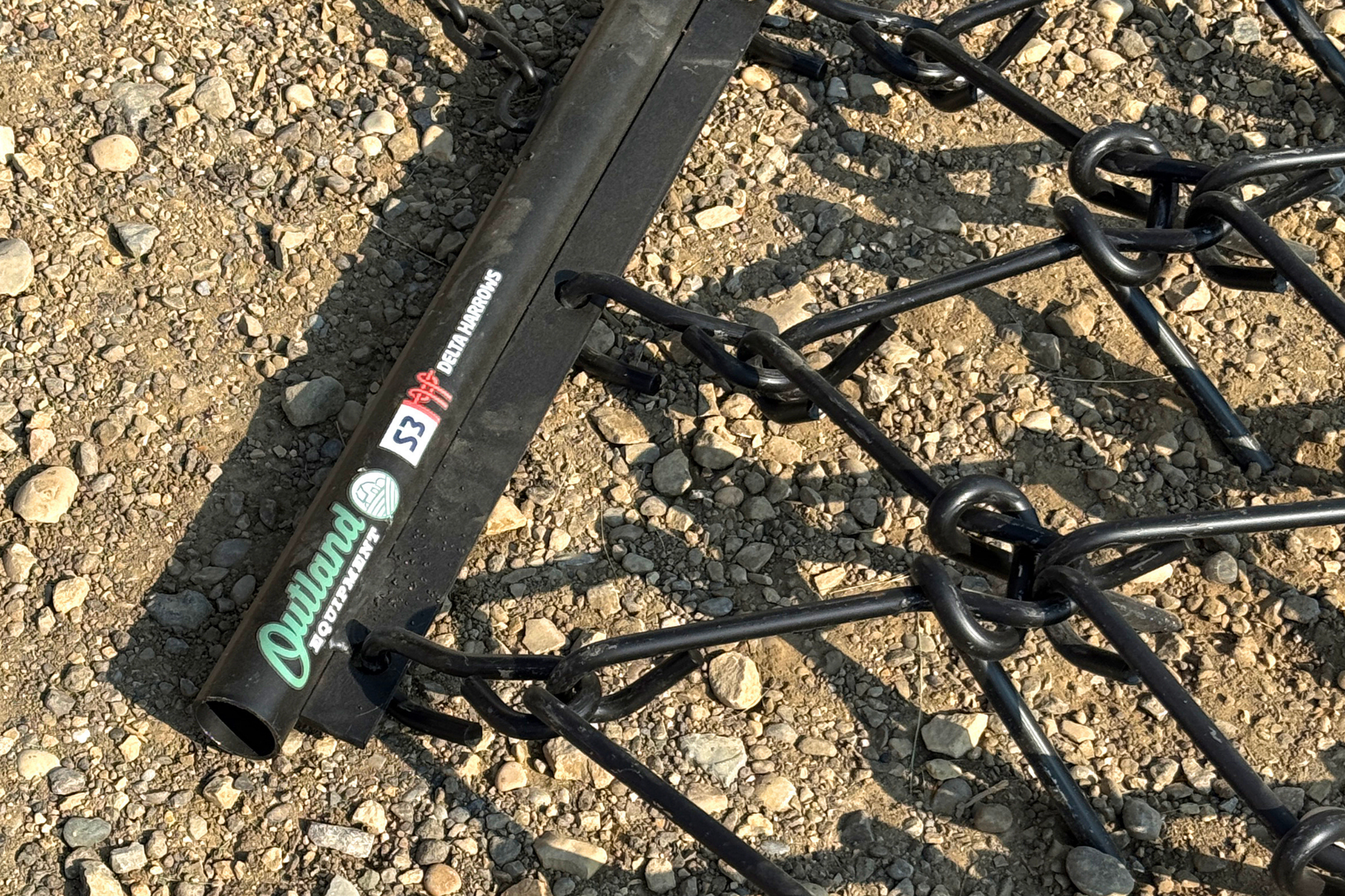 Metal harrow on gravel with decals reading "Outland Equipment" and "S3 Delta Harrows"