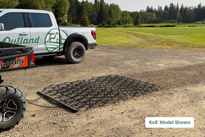 truck with large black harrow in foreground being pulled through gravel on farm