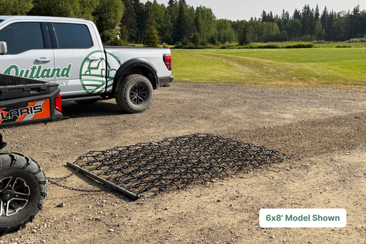 truck with large black harrow in foreground being pulled through gravel on farm